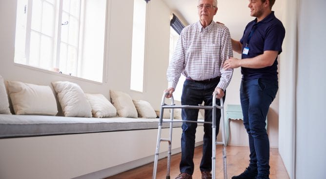 mannelijke verpleger helpt oude man met looprek door gang verpleeghuis